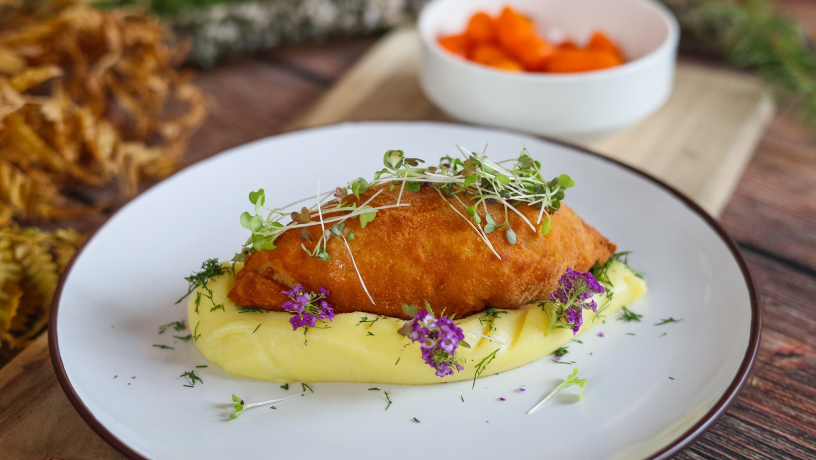 Křupavý řízek na krémovém bramborovém pyré, s mikrolístky a fialovými jedlými květy na bílém talíři