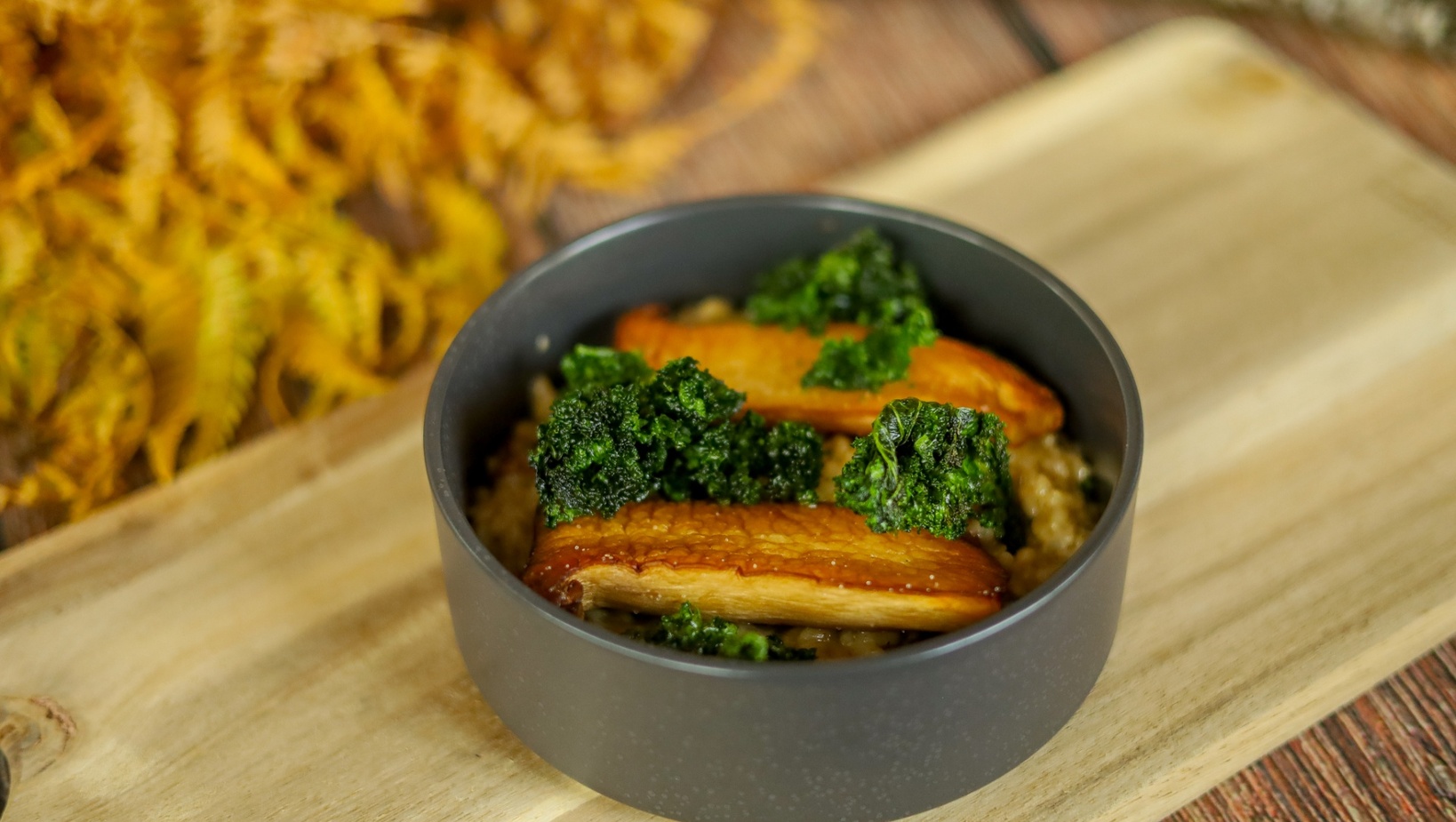 Pečené houby king oyster a křupavý kapustový list na krémovitém rizotu, podávané v tmavé misce na dřevěné desce