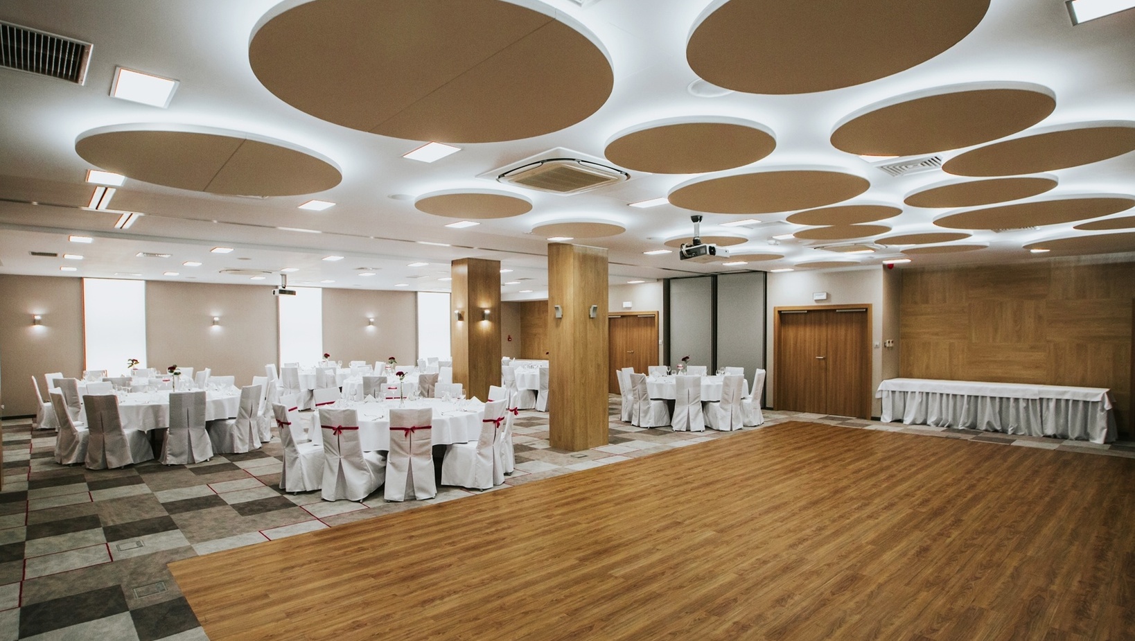 Hotelowa sala bankietowa z drewnianym parkietem, okrągłymi stołami w bieli i krzesłami z czerwonymi kokardami.
