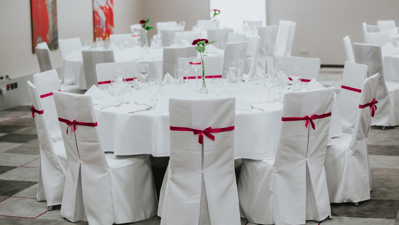 Sala bankietowa z okrągłymi stołami w białych obrusach, krzesła z czerwonymi wstążkami, szkło i pojedyncze czerwone kwiaty.