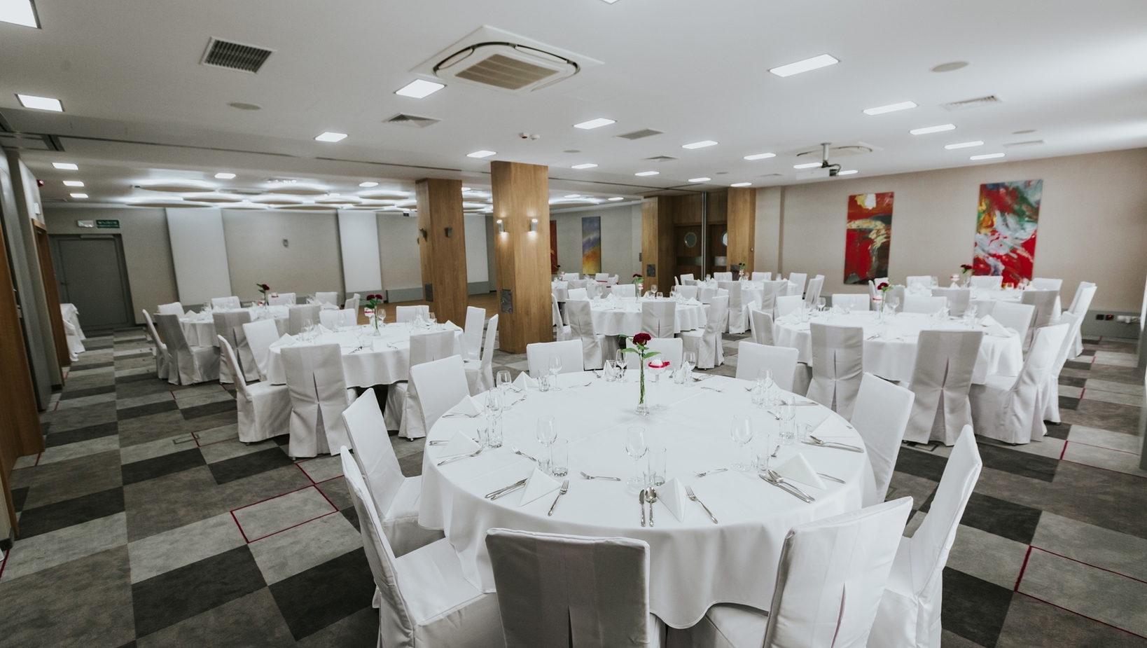 Sala bankietowa z okrągłymi stołami w białych obrusach i pokrowcach, nakryta do uroczystej kolacji z różami.