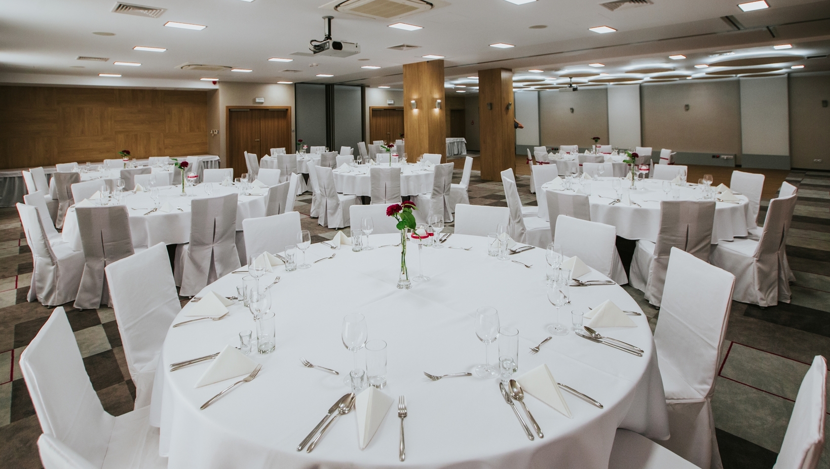 Sala bankietowa z okrągłymi stołami w białych obrusach, pokrytymi krzesłami i czerwonymi różami.