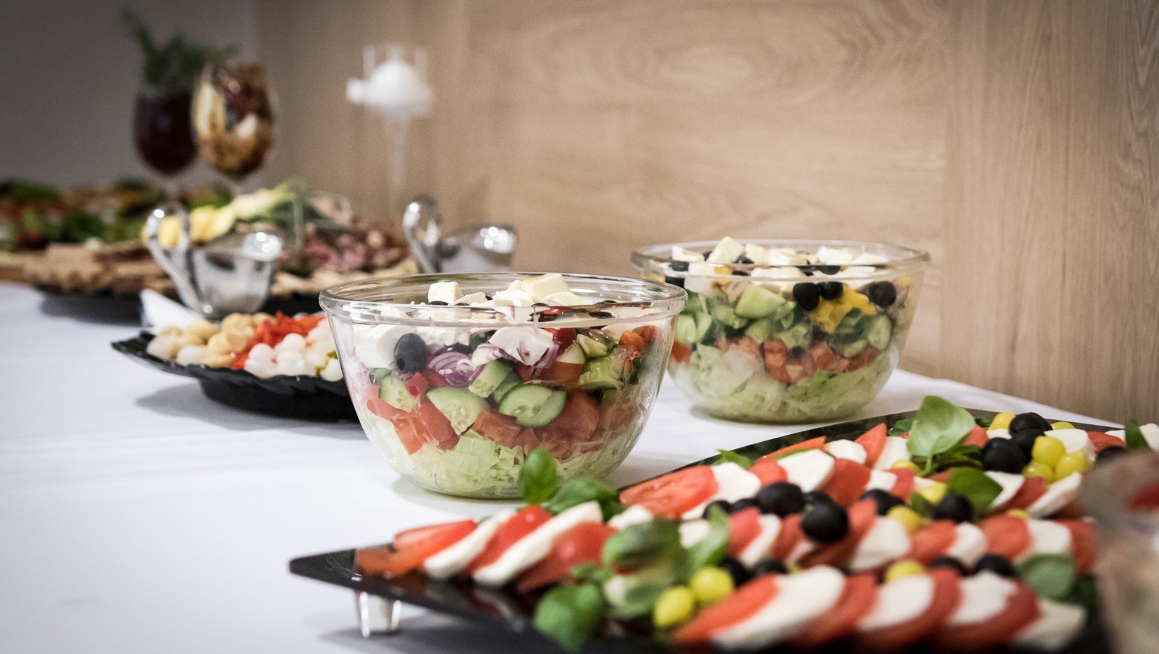 Stół bufetowy ze szklanymi miskami mieszanych sałatek, talerz caprese z pomidorami, mozzarellą i bazylią oraz oliwkami na białym obrusie