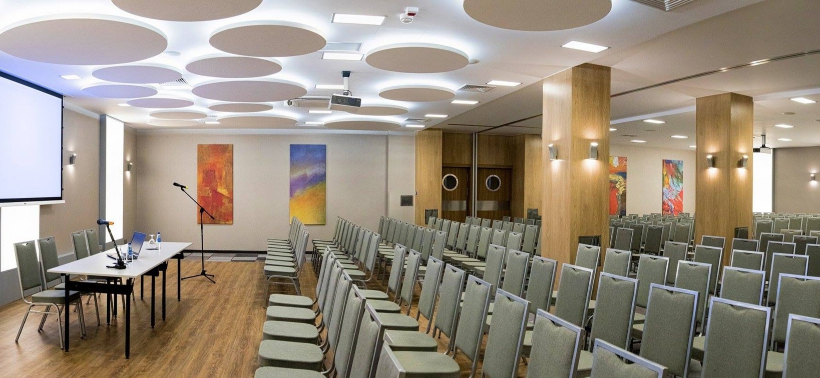 Moderní konferenční síň s řadami židlí, obrazovkou, stolkem pro přednášejícího, kulatými panely a dřevěnými sloupy