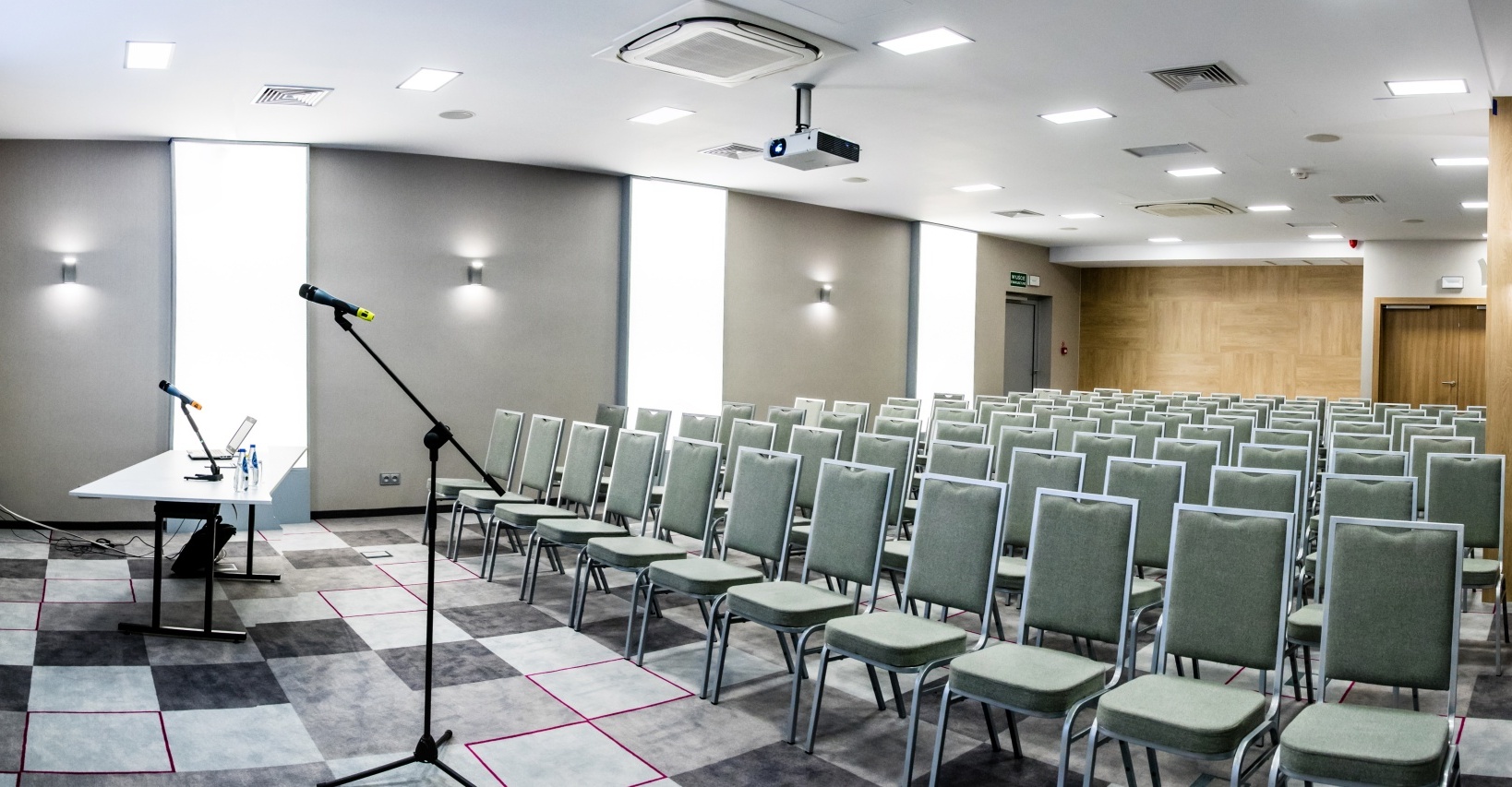 Moderní hotelová konferenční síň s řadami zelených židlí, projektorem a obrazovkou, stolem s notebookem a mikrofony, kobercem a jasným osvětlením