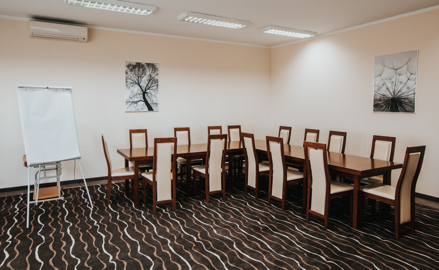 Hotelová konferenční síň s dlouhým dřevěným stolem, krémovými vysokými židlemi, flipchartem a klimatizací.