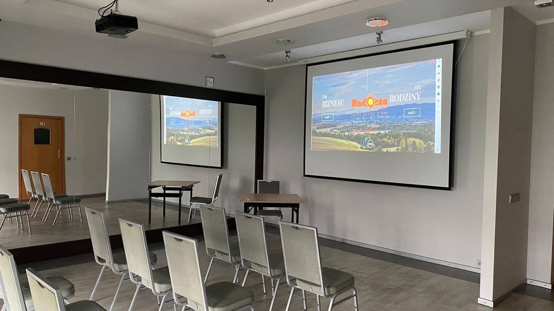 Malá konferenční místnost v hotelu s projektorem na stropě a velkou obrazovkou, řadami čalouněných židlí, zrcadlem a stolky.