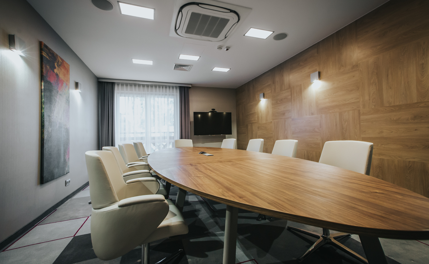 Konferenční místnost v hotelu s oválným dřevěným stolem, bílými koženými židlemi na kolečkách, televizí na stěně a teplým osvětlením