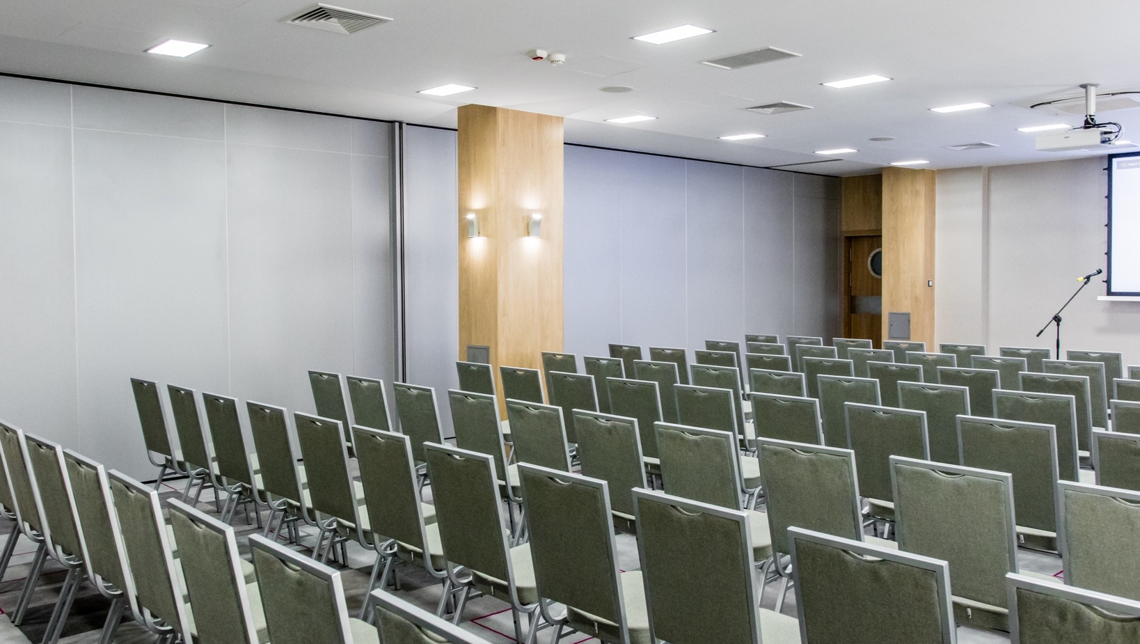 Konferenční místnost v hotelu s řadami zelených židlí, projektorem, mikrofonem, dřevěnými sloupy a jasným osvětlením
