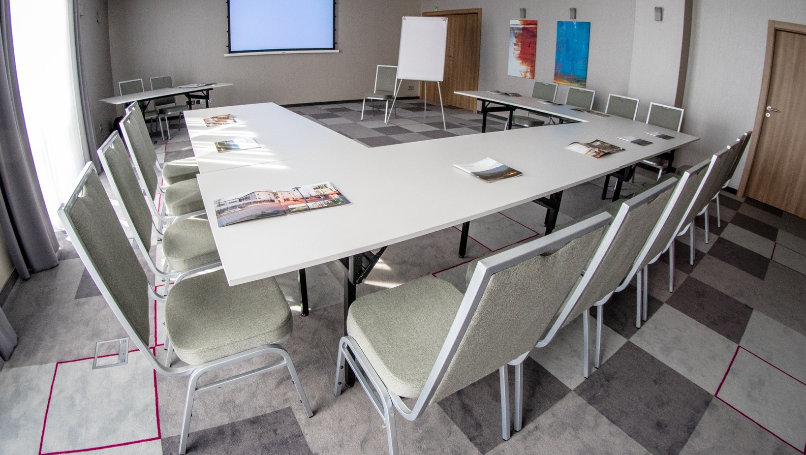 Konferenční místnost v hotelu v uspořádání U: bílé stoly, čalouněné židle, projekční obrazovka, flipchart a brožury.