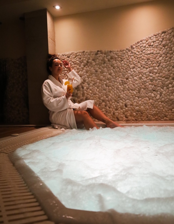 Gość w białym szlafroku relaksuje przy bąbelkowym jacuzzi, trzyma koktajl przy kamiennej ścianie w ciepłym świetle