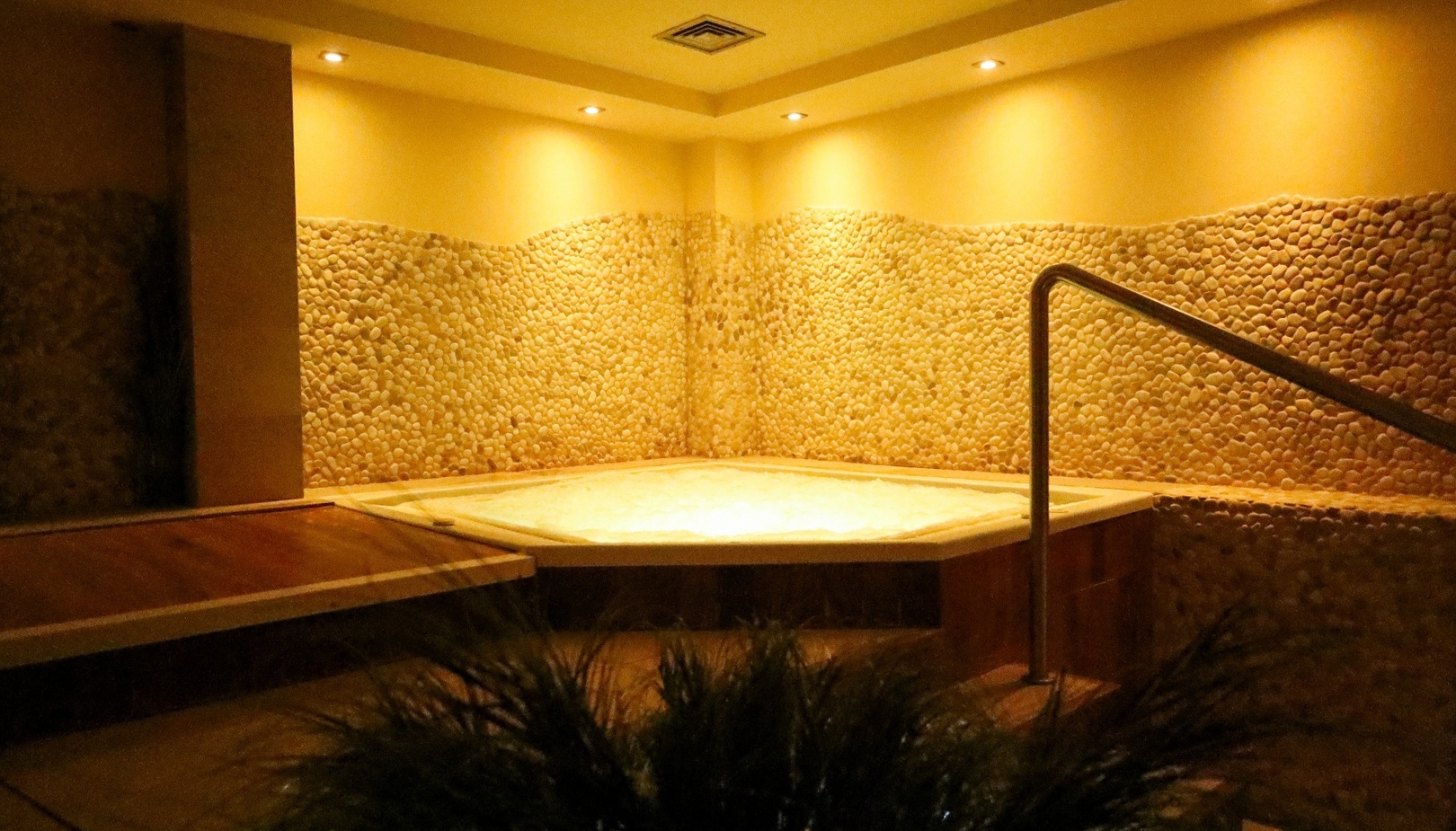 Jacuzzi w spa z ciepłym oświetleniem, ścianami wyłożonymi kamykami, drewnianym tarasem i metalową poręczą.