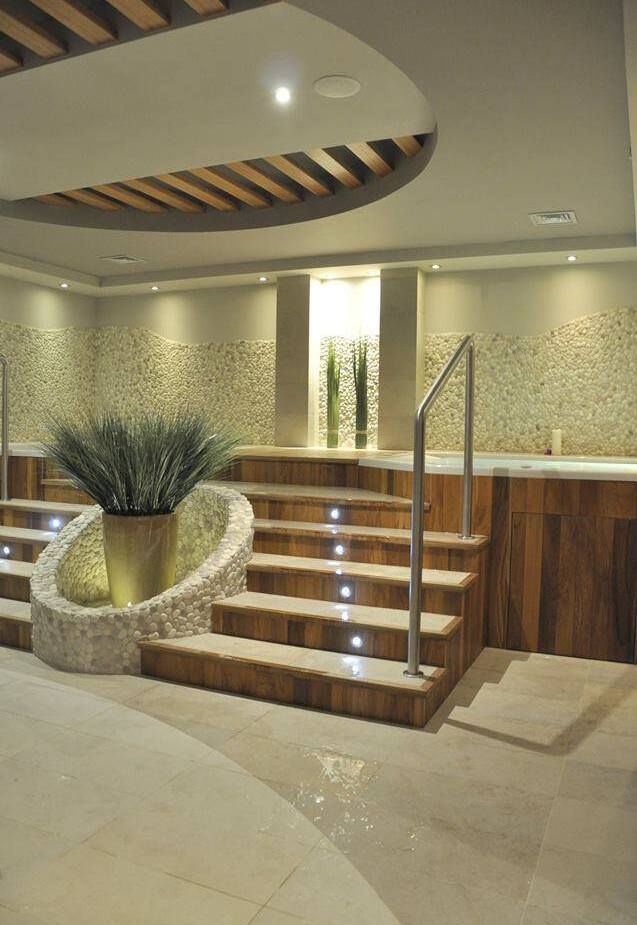 Spa wewnętrzne z podwyższonymi drewnianymi schodami prowadzącymi do wanny z hydromasażem, ściany z kamyków, nastrojowe oświetlenie i rośliny.