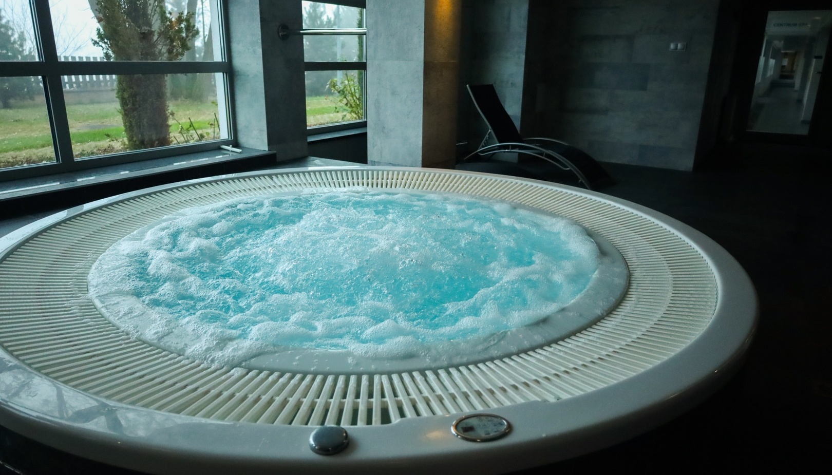 Jacuzzi w hotelowym spa, bąbelki, leżaki przy dużych oknach z widokiem na ogród