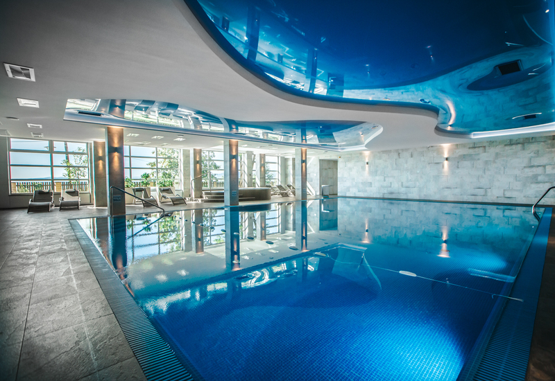 Kryty basen hotelowy z spokojną błękitną wodą, giętym lustrzanym sufitem, dużymi oknami i leżakami na słonecznym tarasie