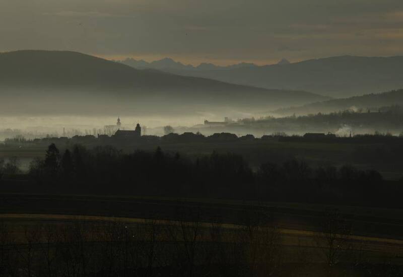 Mglista dolina o świcie z sylwetką wioski i kościelną wieżą, warstwowe wzgórza i odległe szczyty górskie w miękkim świetle