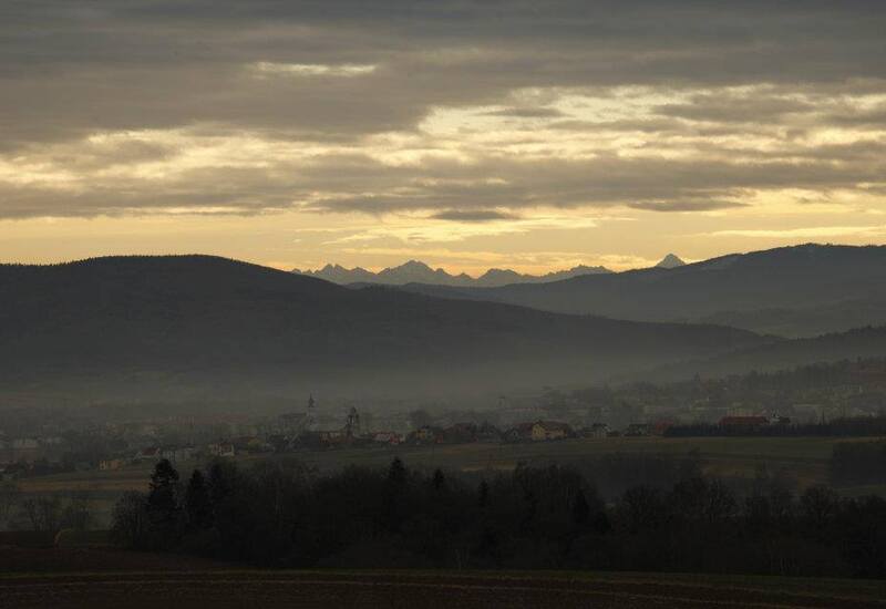 Mglista panorama doliny z tarasu: wieś z wieżami kościoła, falujące wzgórza i odległe szczyty gór o złotym świcie