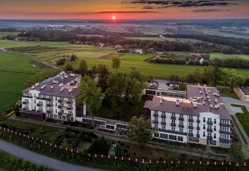 Wiejski hotel z dwoma połączonymi budynkami, balkonami przy pokojach, przeszkloną restauracją, dziedzińcem i wschodem słońca nad polami