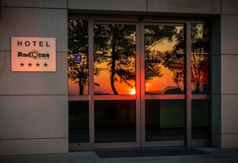 Wejście do hotelu z przeszklonymi drzwiami odbijającymi ciepły zachód słońca i drzewa; tablica z czterema gwiazdkami i symbolem dostępności.