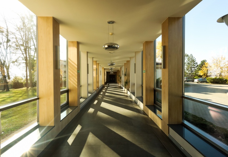Długi hotelowy korytarz z oknami od podłogi do sufitu, drewnianymi filarami, wiszącymi lampami i siedziskami, z widokiem na ogród i parking.