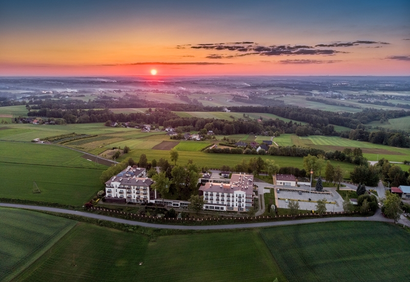 Widok z lotu ptaka na hotel na wsi z balkonami, parkingiem i ogrodami pośród falistych pól o wschodzie słońca