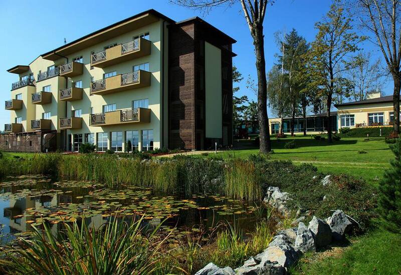 Czteropiętrowy hotel nad stawem z balkonami, liliami wodnymi, zadbanym trawnikiem i dojrzałymi drzewami.