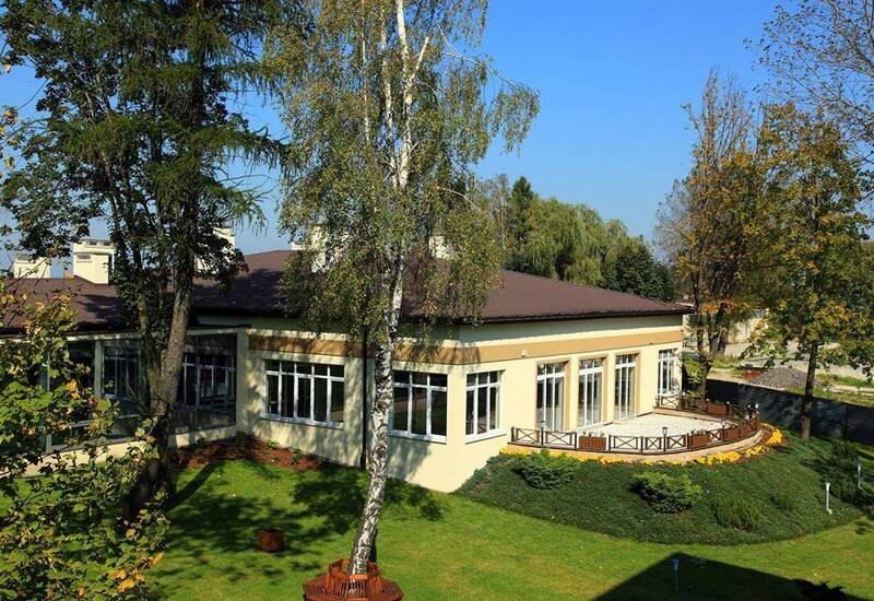 Parterowa sala restauracyjna z dużymi oknami i zaokrąglonym tarasem, otoczona trawnikiem, drzewami i rabatami