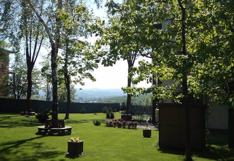 Słoneczny ogród hotelowy z zadbaną trawą, drzewami, okrągłą ławką i widokiem na odległe wzgórza