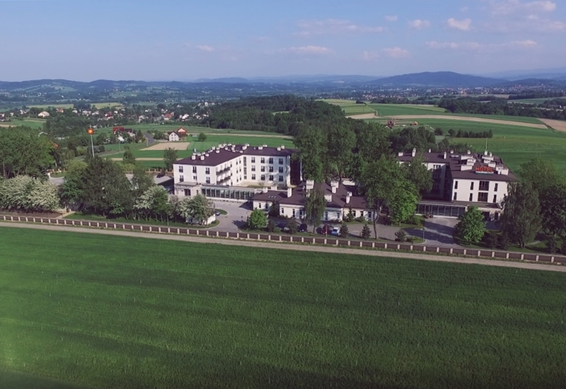 Trzypiętrowy biały hotel z przeszklonym tarasem, zadrzewionym dziedzińcem i parkingiem, otoczony zielonymi polami i wzgórzami.