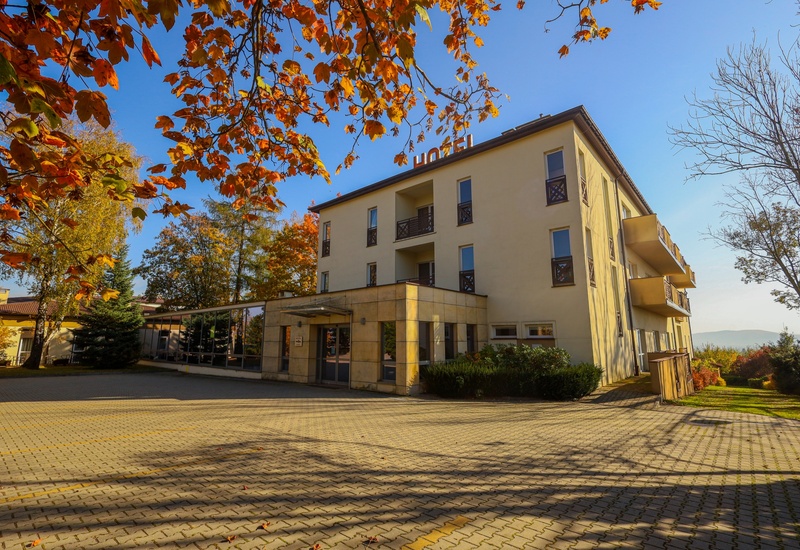 Trzykondygnacyjny hotel z balkonami, przeszklonym wejściem, jesiennymi drzewami i wybrukowanym, przestronnym parkingiem