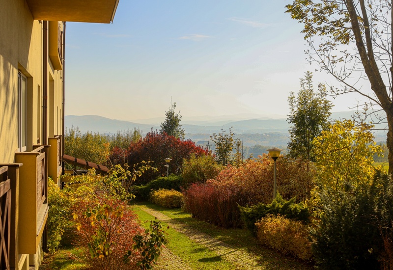 Żółty hotel z drewnianymi balkonami, zadbaną alejką w ogrodzie, jesienne krzewy i latarnie, widok na pofałdowane wzgórza.