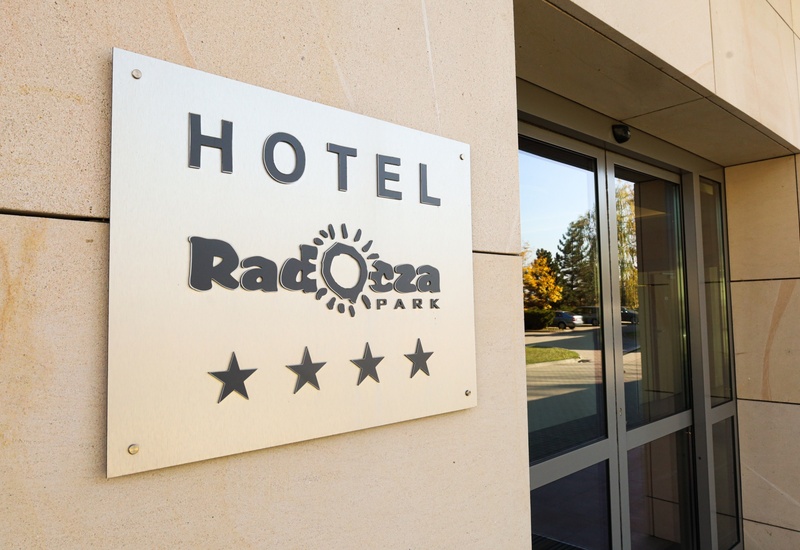 Tablica z nazwą czterogwiazdkowego hotelu na piaskowcowej elewacji obok szklanych drzwi wejściowych i parkingu