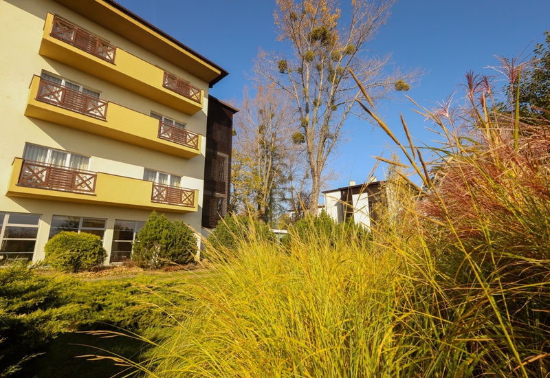 Trzypiętrowy hotel z pokojami i drewnianymi balkonami, widok na zadbany jesienny ogród, wysokie drzewa i błękitne niebo.