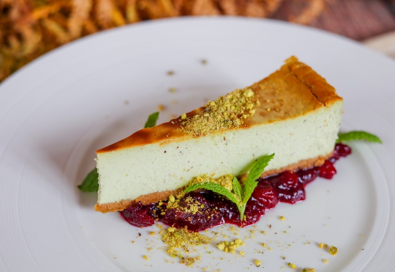 Kawałek sernika pistacjowego z pokruszonymi pistacjami, czerwonym sosem owocowym i miętą na białym talerzu