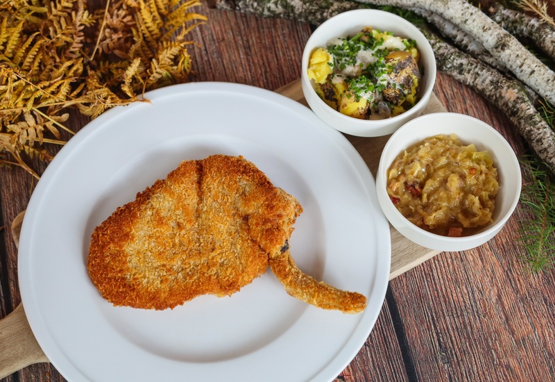 Chrupiący panierowany kotlet z kością, podany z przypieczonymi masłowymi ziemniakami i kremową duszoną kapustą.