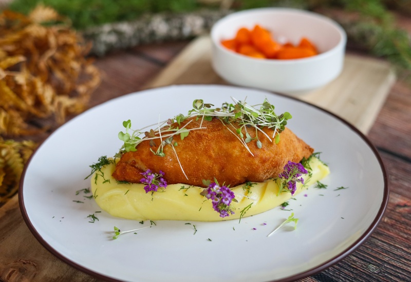 Chrupiący panierowany filet na kremowym puree ziemniaczanym, z mikroliśćmi i fioletowymi jadalnymi kwiatami na białym talerzu