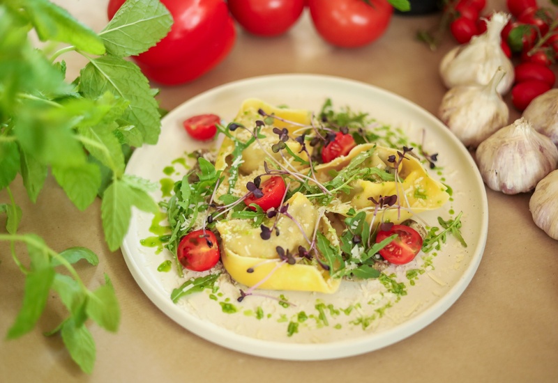 Świeże nadziewane ravioli z pomidorkami koktajlowymi, rukolą, kiełkami i startym serem skropione olejem ziołowym na białym talerzu