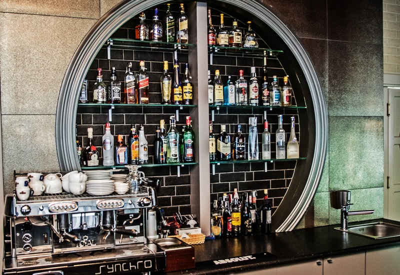 Okrągła podświetlana półka barowa z butelkami, ekspres do kawy z filiżankami na blacie, czarna kafelkowa ściana i zlew