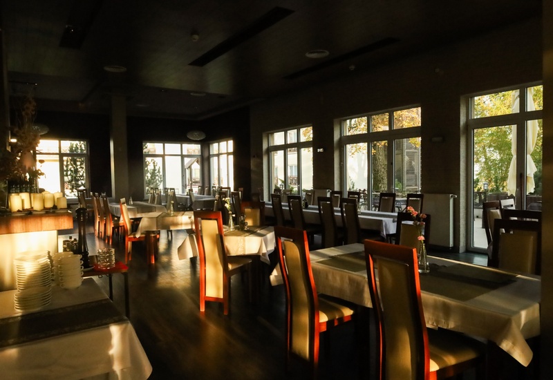 Sala restauracyjna hotelu z białymi obrusami, drewnianymi krzesłami, wazonikami z kwiatami i złotym światłem przez duże okna