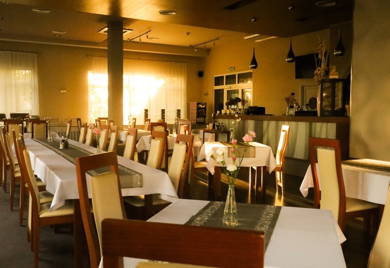 Restauracja hotelowa z długimi stołami, białymi obrusami, drewnianymi krzesłami, wazonikami z kwiatami, ciepłym światłem i barem.