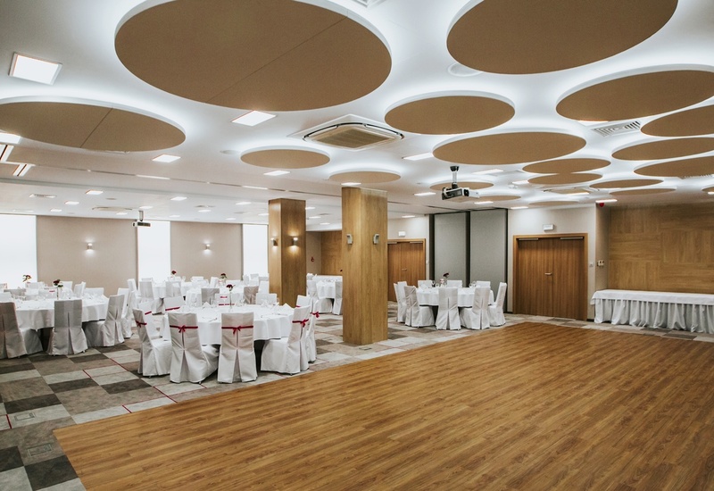 Hotelowa sala bankietowa z drewnianym parkietem, okrągłymi stołami w bieli i krzesłami z czerwonymi kokardami.