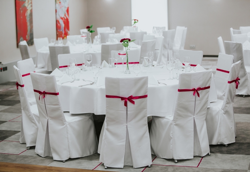 Sala bankietowa z okrągłymi stołami w białych obrusach, krzesła z czerwonymi wstążkami, szkło i pojedyncze czerwone kwiaty.