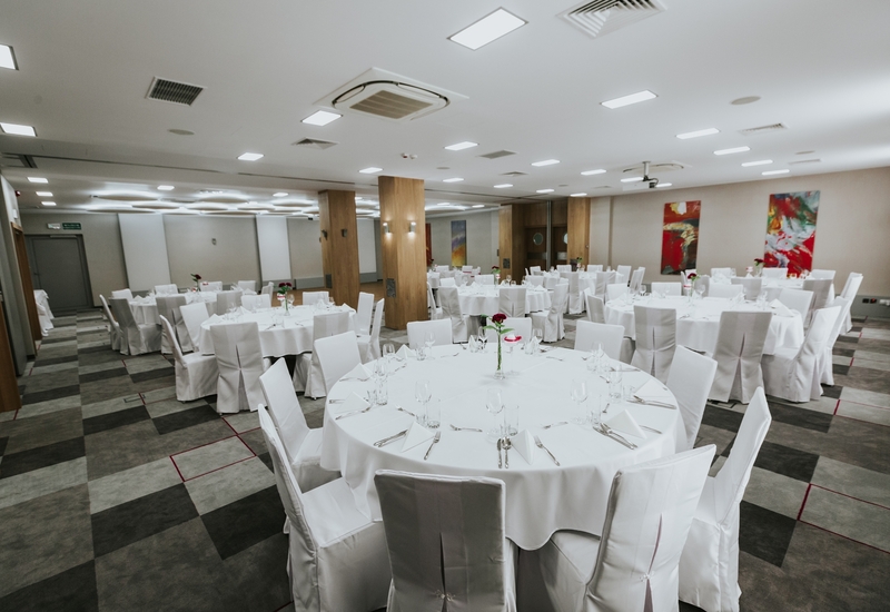 Sala bankietowa z okrągłymi stołami w białych obrusach i pokrowcach, nakryta do uroczystej kolacji z różami.