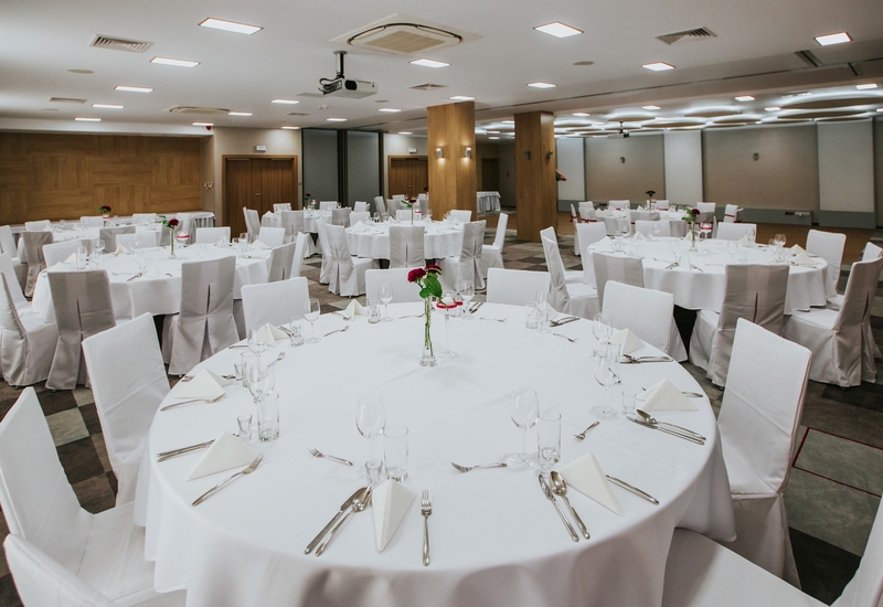 Sala bankietowa z okrągłymi stołami w białych obrusach, pokrytymi krzesłami i czerwonymi różami.