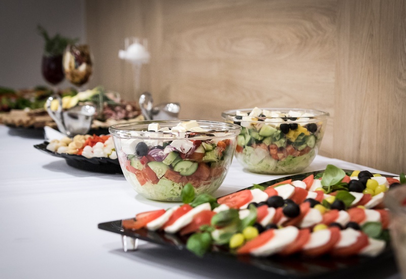Stół bufetowy ze szklanymi miskami mieszanych sałatek, talerz caprese z pomidorami, mozzarellą i bazylią oraz oliwkami na białym obrusie