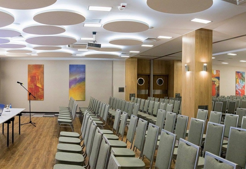 Nowoczesna sala konferencyjna z rzędami krzeseł, ekranem, stolikiem prelegenta, okrągłymi panelami i drewnianymi słupami