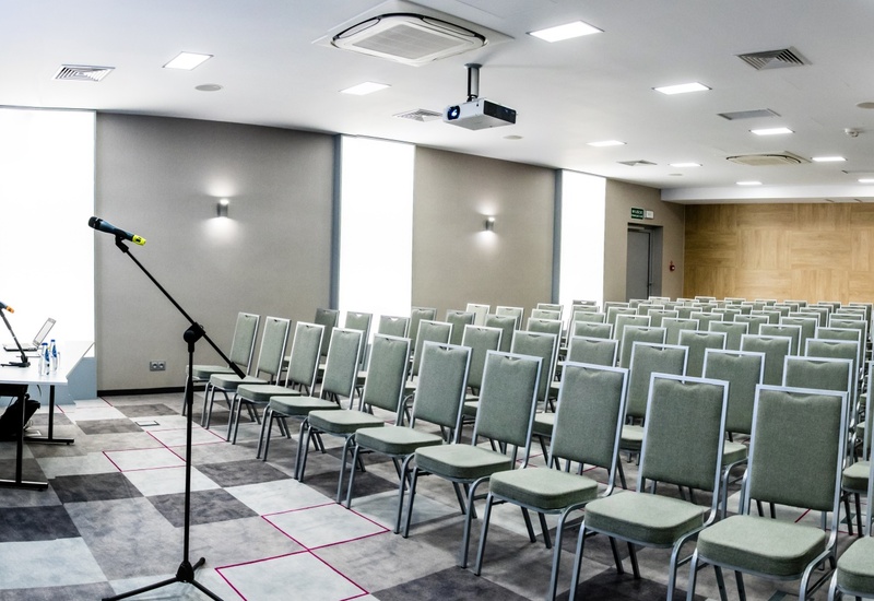 Nowoczesna sala konferencyjna w hotelu z rzędami zielonych krzeseł, projektorem i ekranem, stołem z laptopem i mikrofonami, wykładzina i jasne oświetlenie
