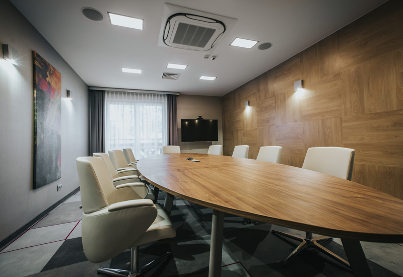 Sala konferencyjna w hotelu z owalnym drewnianym stołem, białymi skórzanymi krzesłami na kółkach, telewizorem na ścianie i ciepłym oświetleniem
