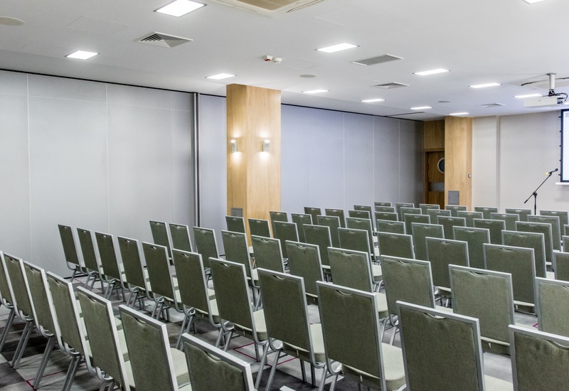 Sala konferencyjna w hotelu z rzędami zielonych krzeseł, projektorem, mikrofonem, drewnianymi filarami i jasnym oświetleniem