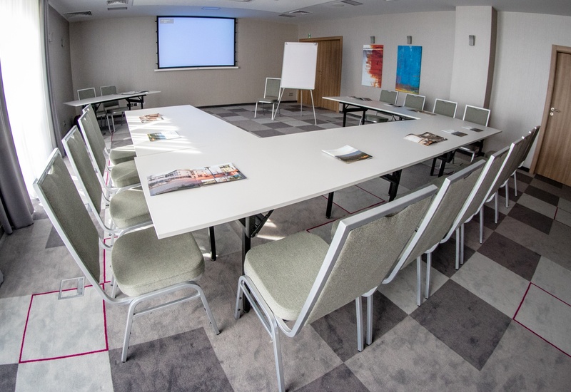 Sala konferencyjna w hotelu w układzie U: białe stoły, tapicerowane krzesła, ekran projektora, flipchart i broszury.